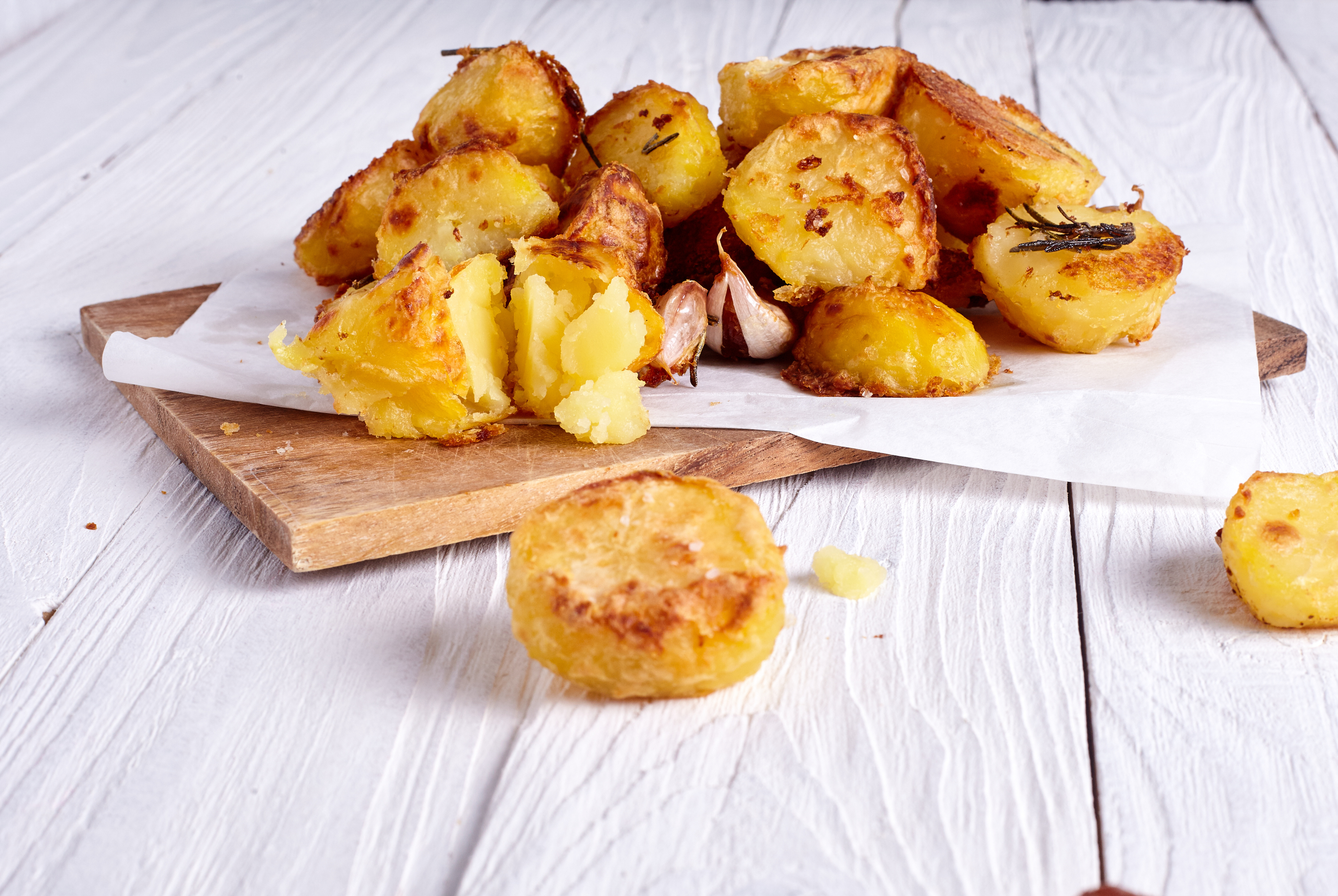 Crispy roast potatoes with fluffy middles, garlic cloves and rosemary, served on parchment over a wooden board.